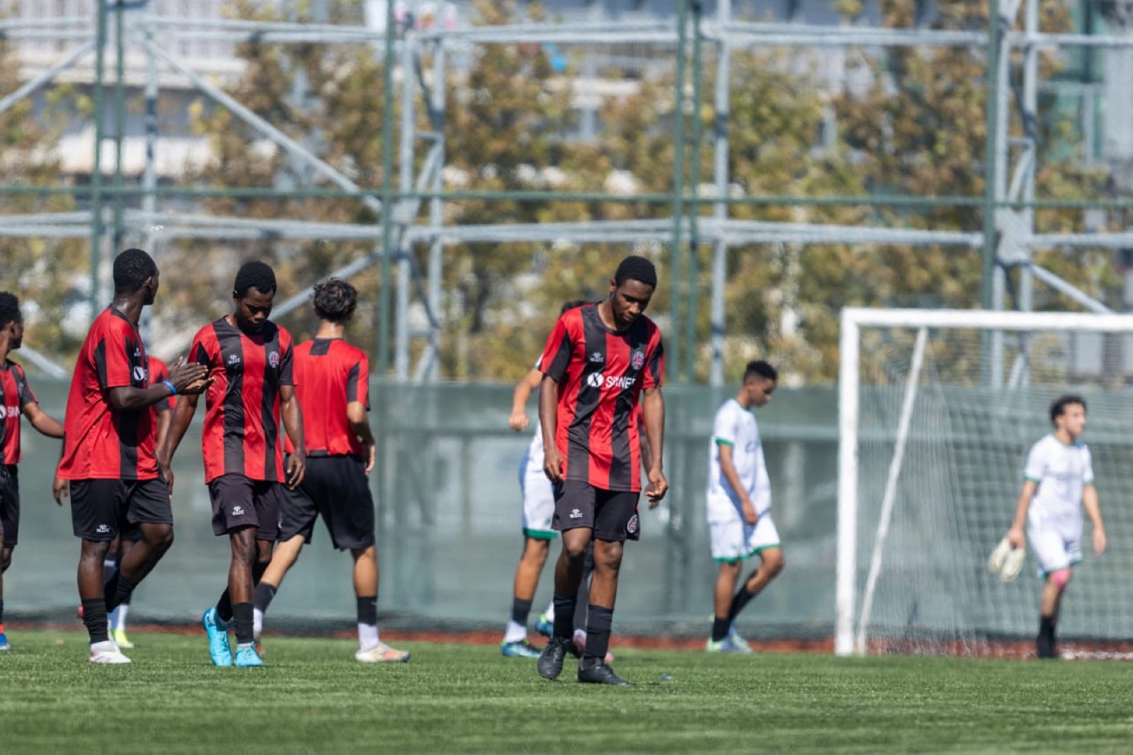 Football players in red and black kits beside a goal on the pitch.